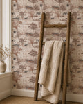 Rustic countryside toile wallpaper with barns and trees in sepia tones, wooden ladder with beige blanket.